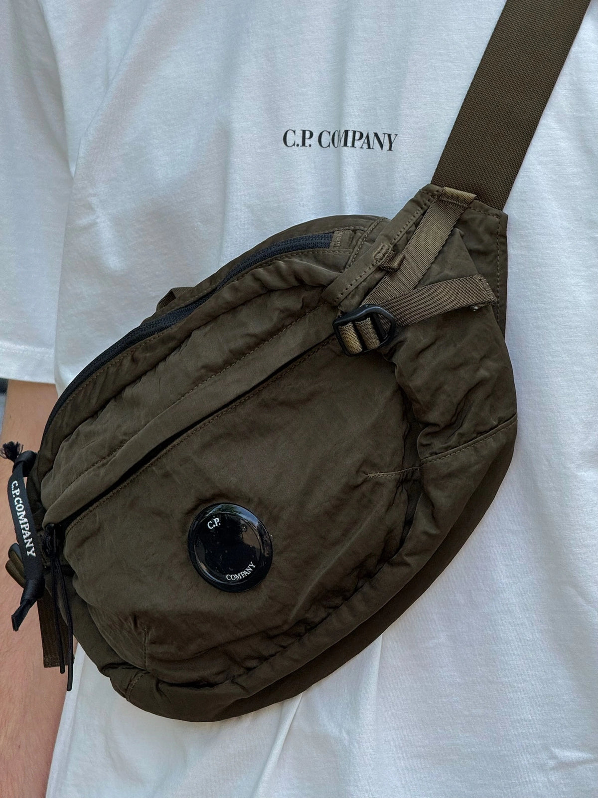 An olive green C.P. Company crossbody bag with a black circular logo patch, worn across a white t-shirt.