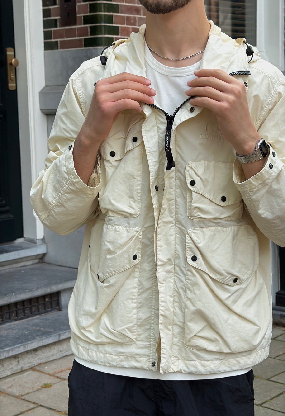 A man wearing a cream-colored lightweight utility jacket over a white t-shirt, standing outdoors in front of a building entrance.