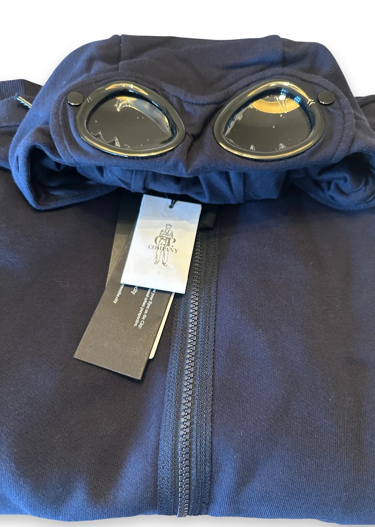 A folded navy blue fleece jacket with a hood featuring two circular goggles, shown with retail tags on a white background.