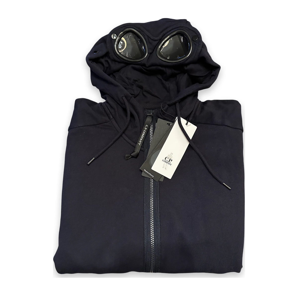 A folded black hooded sweatshirt with a full front zipper, black drawstrings, and a pair of round black goggles attached to the hood, shown on a white background with retail tags attached.