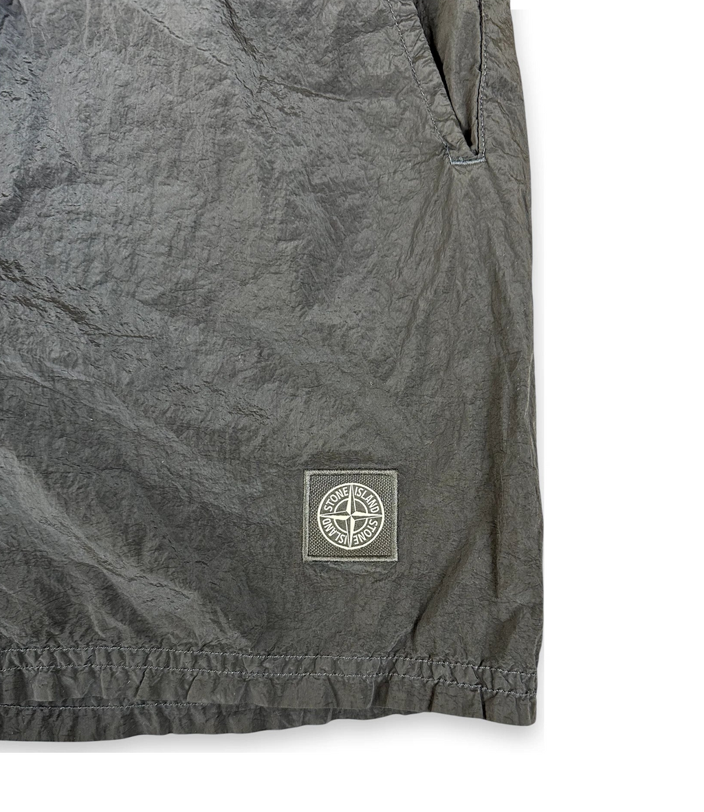 Close-up of dark gray crinkled fabric with a small square Stone Island logo patch near the lower right area, on a white background.