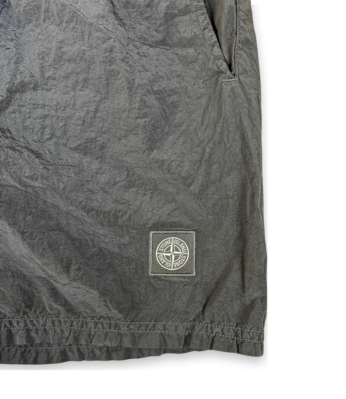 Close-up of dark gray crinkled fabric with a small square Stone Island logo patch near the lower right area, on a white background.