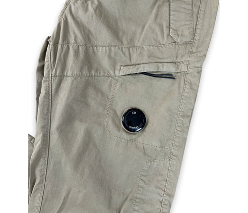 Close-up of a pair of khaki cargo pants showing a side pocket with a black button closure and a zippered pocket above it, on a white background.