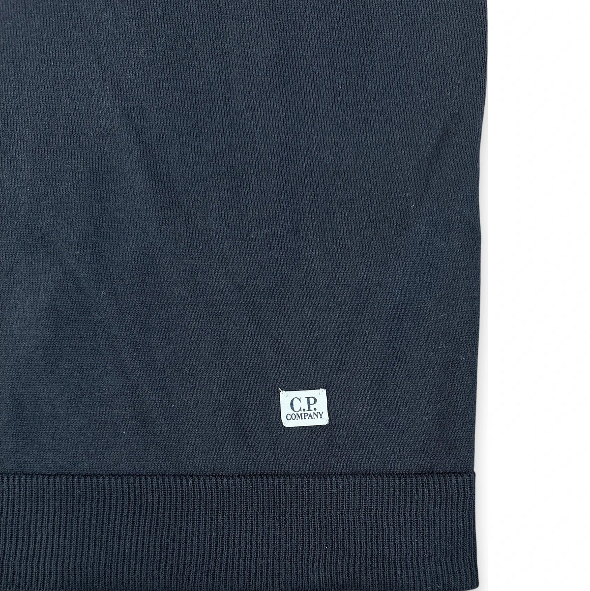 Close-up of a dark navy ribbed knit garment, likely a sweater or sweatshirt, showing a small white C.P.