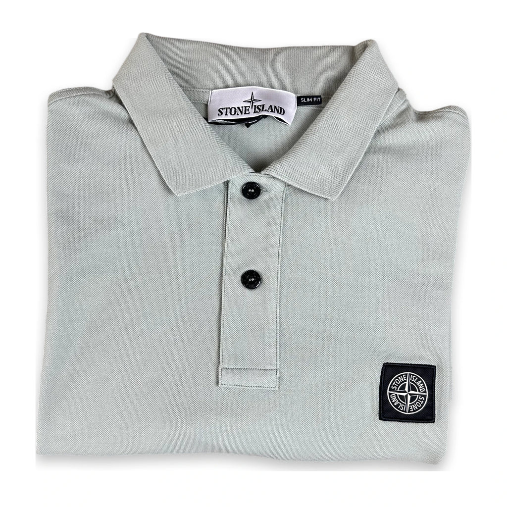 A folded Stone Island polo shirt in light gray, featuring a classic collar, two black buttons, and the signature compass logo patch on the left chest.