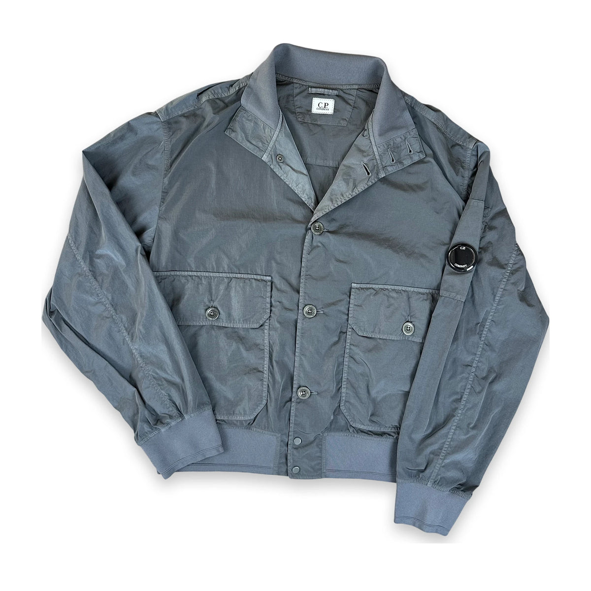 A gray lightweight jacket with a button-up front, two front flap pockets, and a circular black patch on the left sleeve, laid flat on a white background.