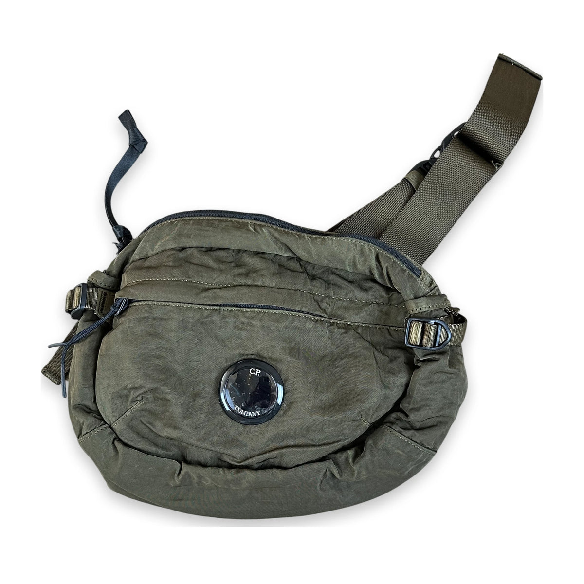 An olive green waxed canvas crossbody bag with a wide adjustable shoulder strap, multiple zippered pockets, and a circular black logo patch on the front, shown against a plain white background.