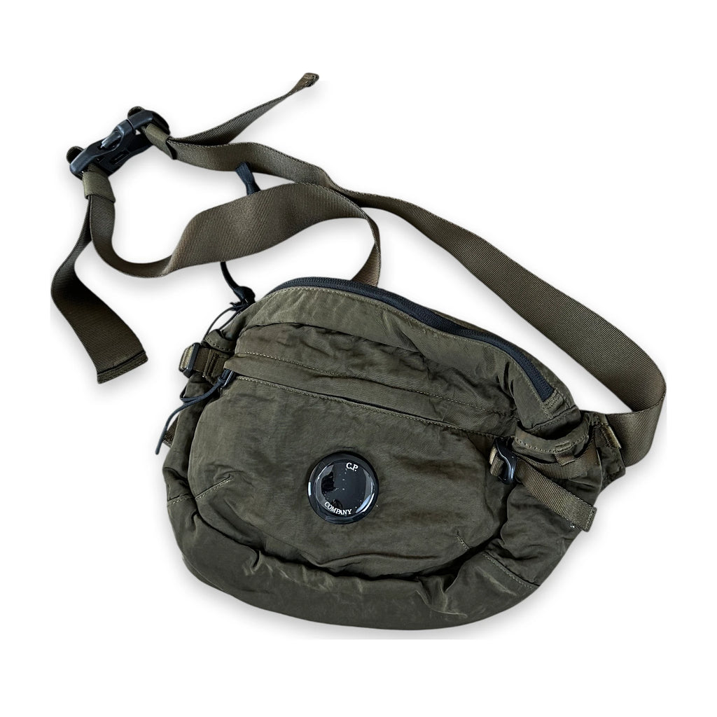 An olive green tactical fanny pack with an adjustable webbing strap and a circular black valve on the front pocket, shown against a plain white background.