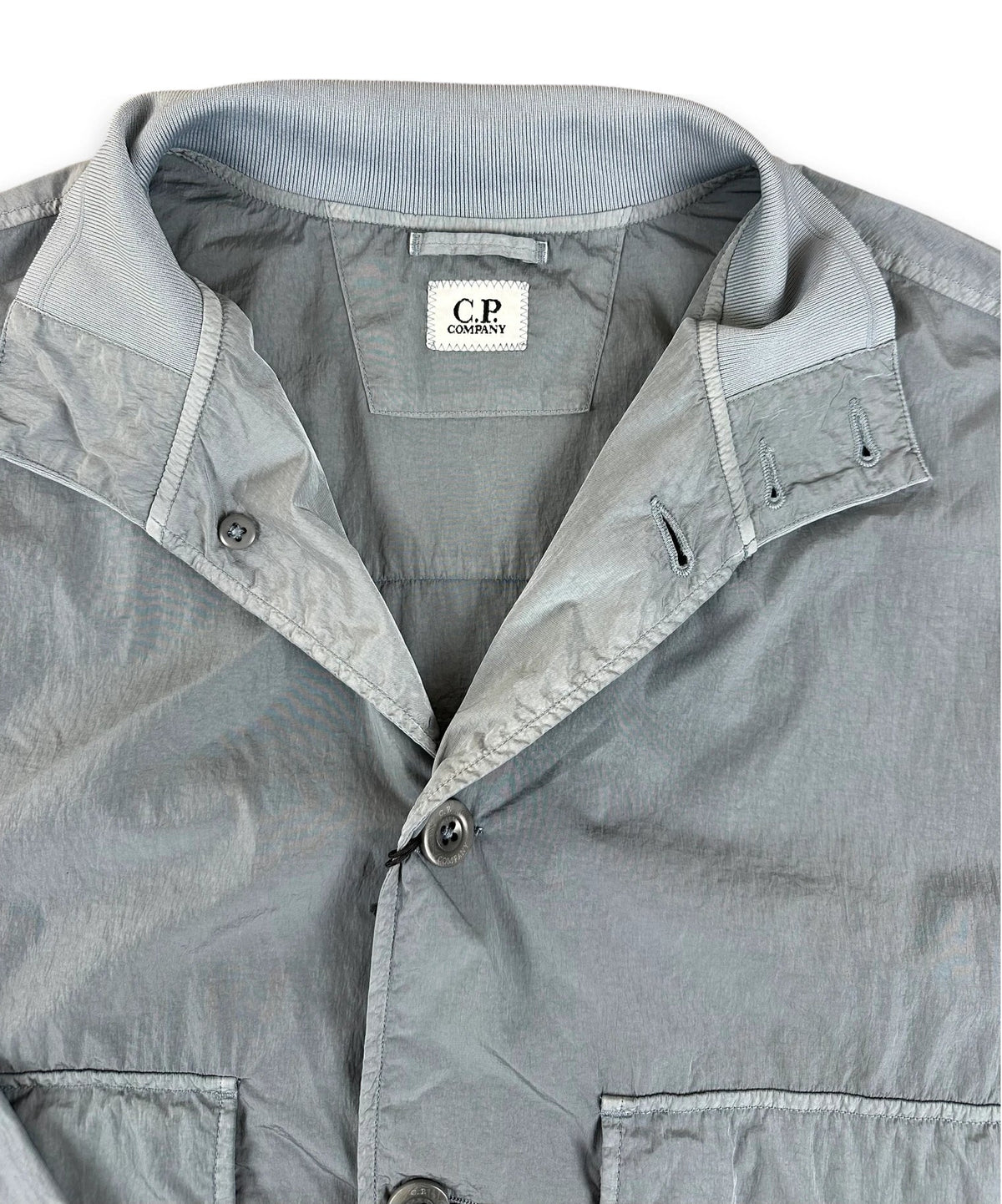 A close-up of a light gray lightweight jacket or overshirt with a pointed collar, button front, and visible CP Company neck label.