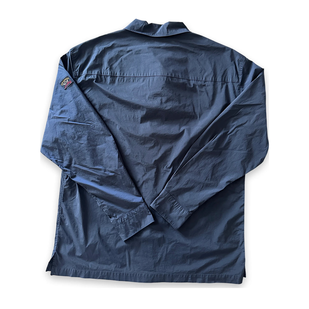 A dark navy blue long-sleeve shirt jacket shown from the back, laid flat on a white background. It features a collar, a vertical seam running down the center, and a small embroidered patch on the left shoulder.