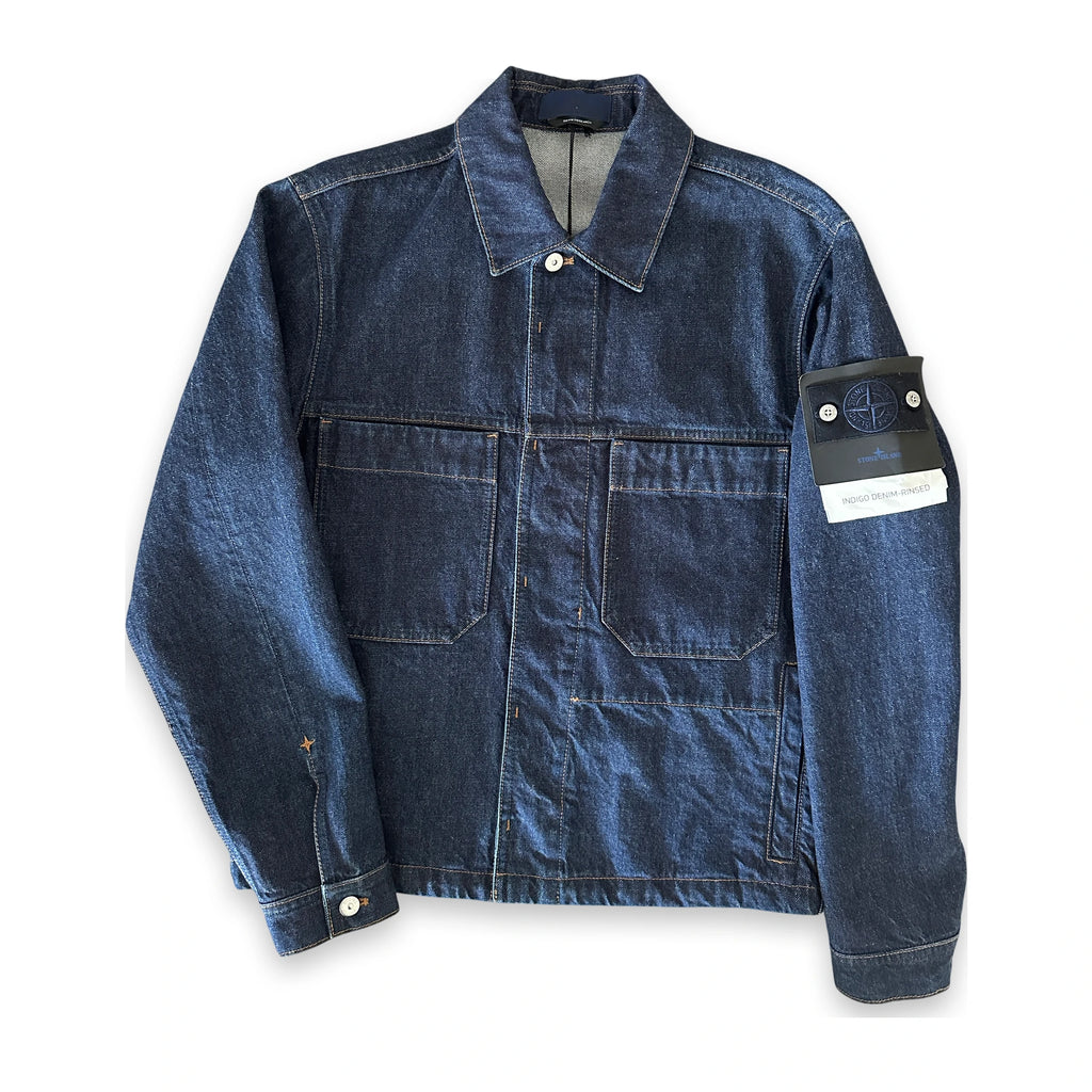 A dark blue denim utility jacket with a collar, button front, and two large chest pockets. The right sleeve features a black Stone Island brand patch with the signature compass logo and a white paper tag attached.