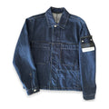 A dark blue denim utility jacket with a collar, button front, and two large chest pockets. The right sleeve features a black Stone Island brand patch with the signature compass logo and a white paper tag attached.