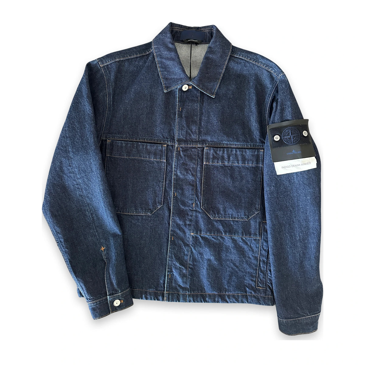 A dark blue denim utility jacket with a collar, button front, and two large chest pockets. The right sleeve features a black Stone Island brand patch with the signature compass logo and a white paper tag attached.