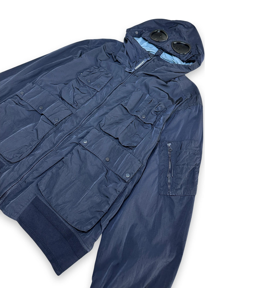 A navy blue hooded utility jacket with multiple front flap pockets and a zippered sleeve, laid flat on a white background. A pair of round sunglasses rests on the hood.