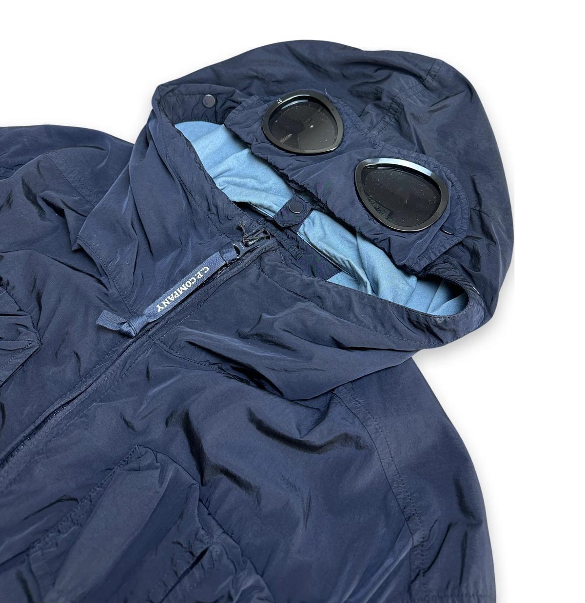 A close-up of a navy blue hooded jacket with a light blue inner lining and two circular black visor-like details on the hood. A small fabric tag on the zipper reads 'CP COMPANY'.
