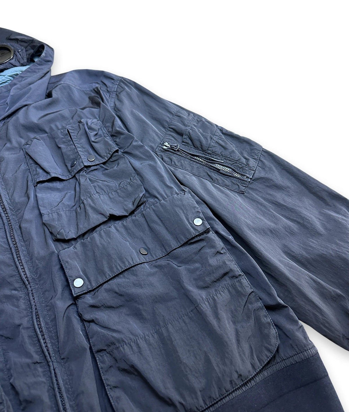 A close-up of a dark navy blue lightweight jacket with multiple front flap pockets and a zippered pocket on the sleeve, laid flat on a white background.
