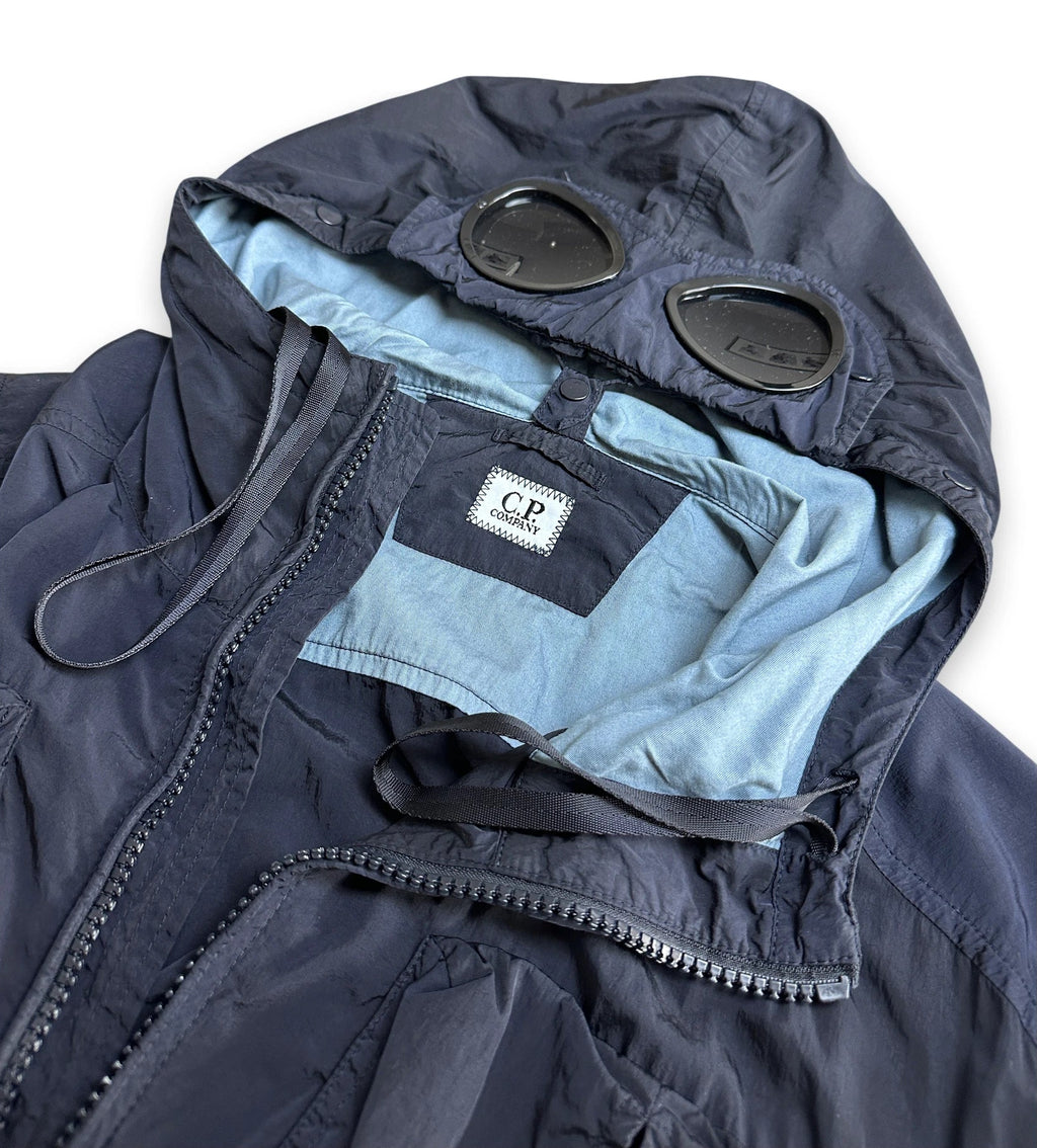 A close-up of a navy blue hooded jacket with a light blue inner lining, featuring a black patch with 'CP Company' on the inside neck label and black drawstrings.