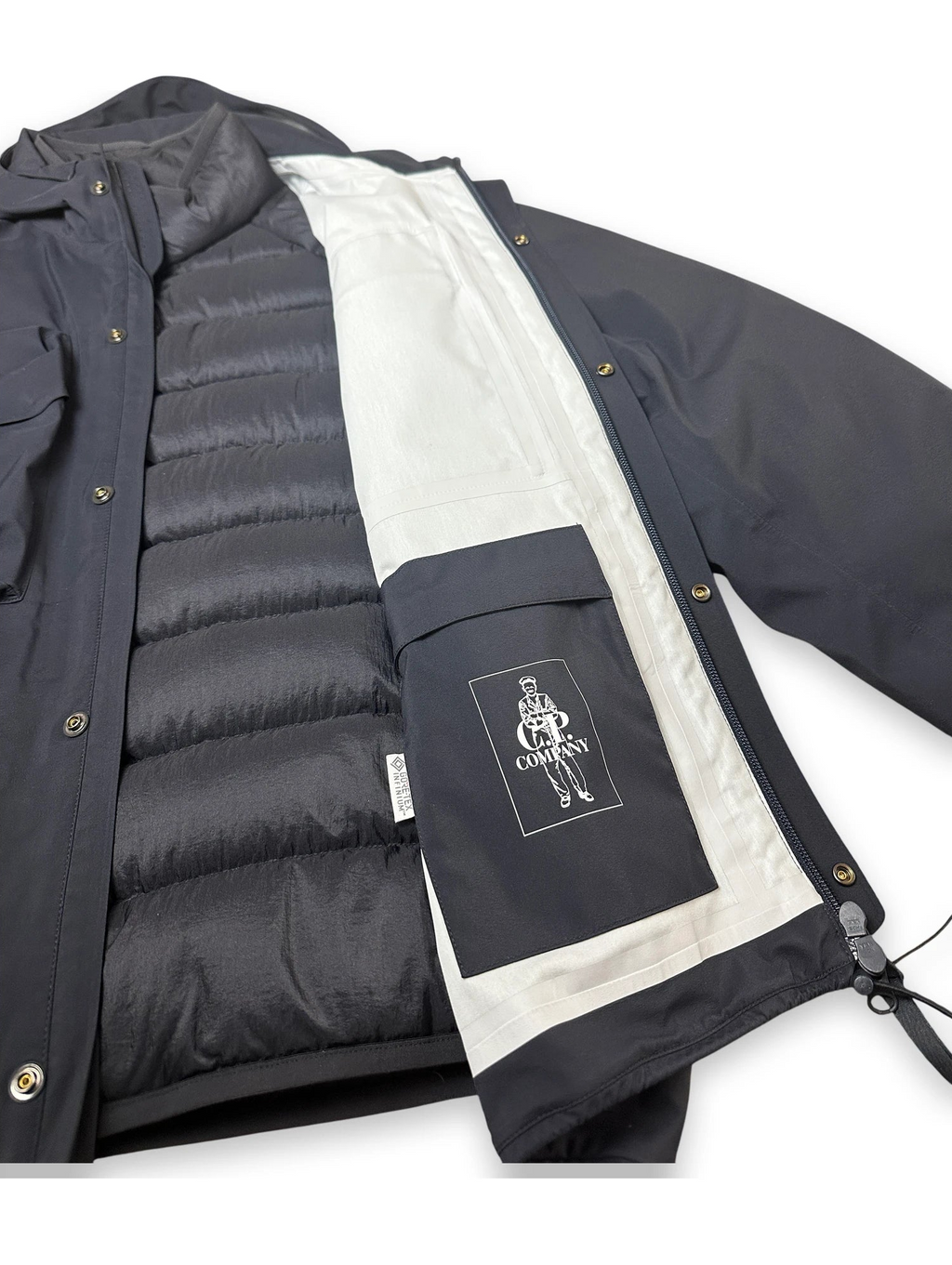 A dark gray outdoor jacket with a black puffer lining and a black front pocket featuring a white 'CP COMPANY' logo patch, shown open on a white background.