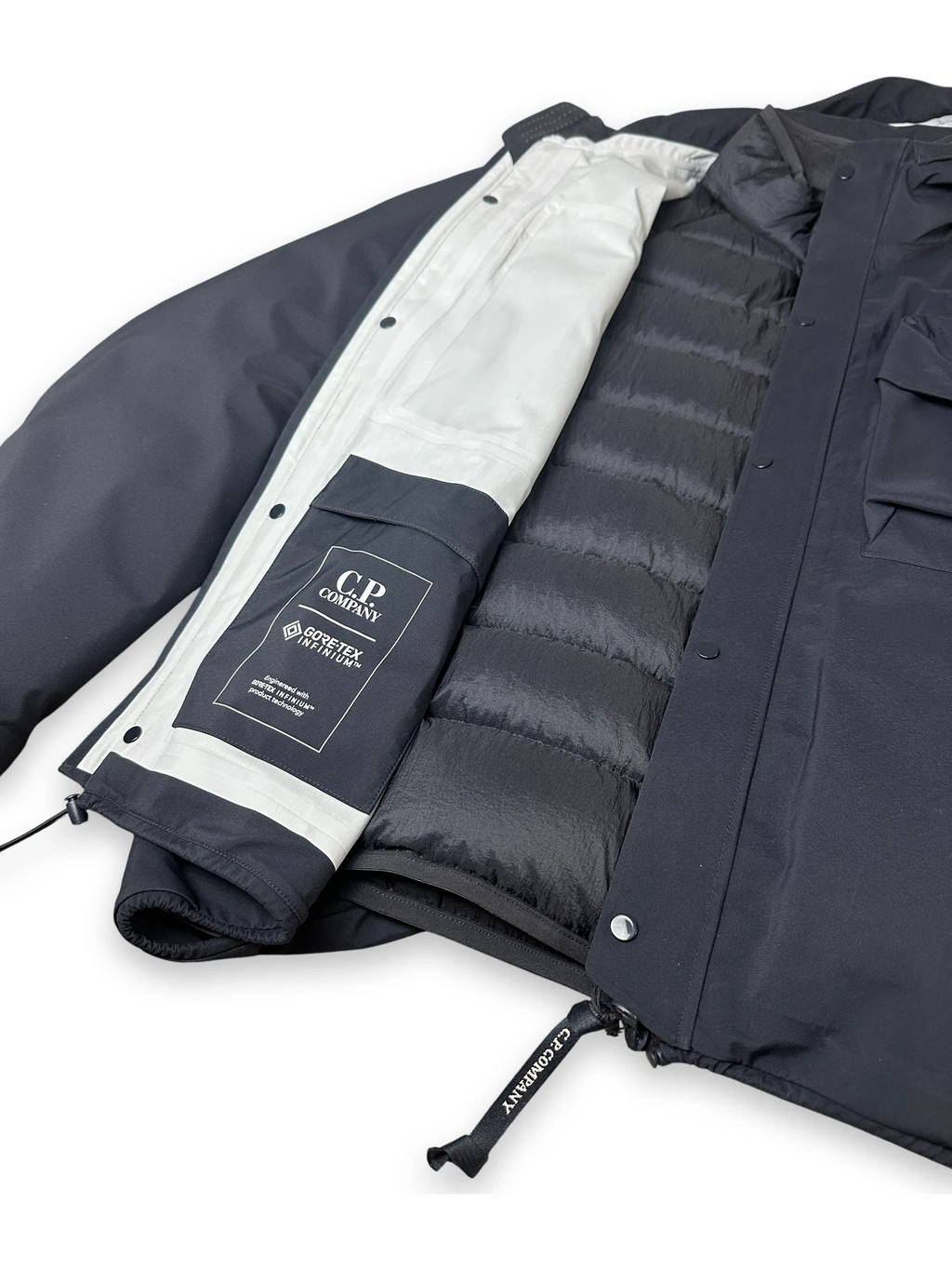 A close-up of a dark navy jacket with its front flap open, showing a white inner lining and a black quilted puffer insert. A black patch on the sleeve reads 'C.P. Company'
