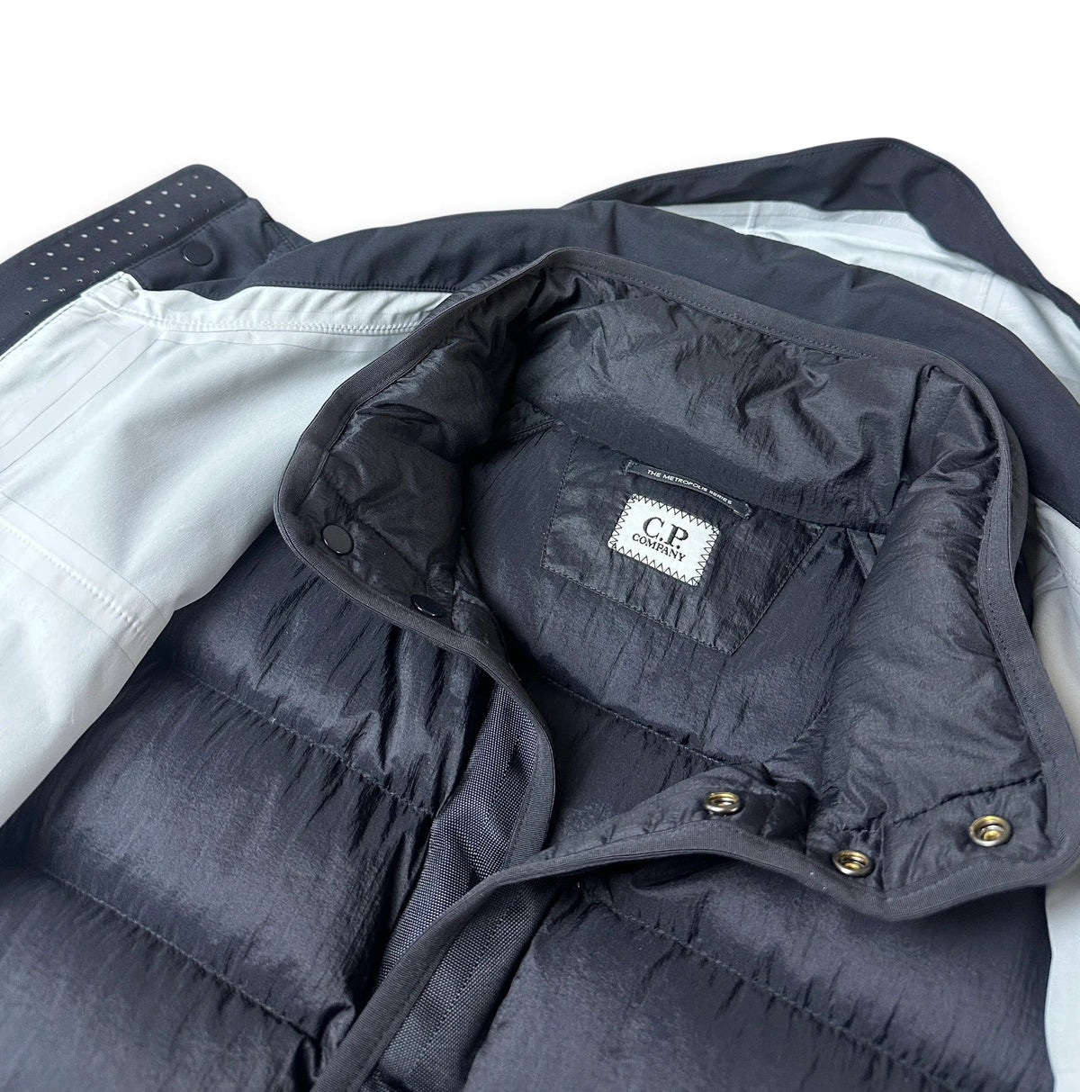 Close-up of a dark navy puffer vest with a light gray lining and a visible 'CP Company' brand label inside the collar.