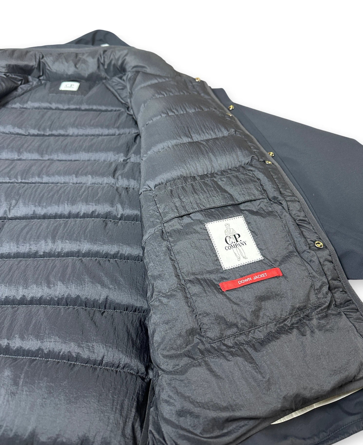 A close-up of a dark gray puffer jacket showing the quilted lining, a white CP Company brand patch, and a red zipper pull on a white background.