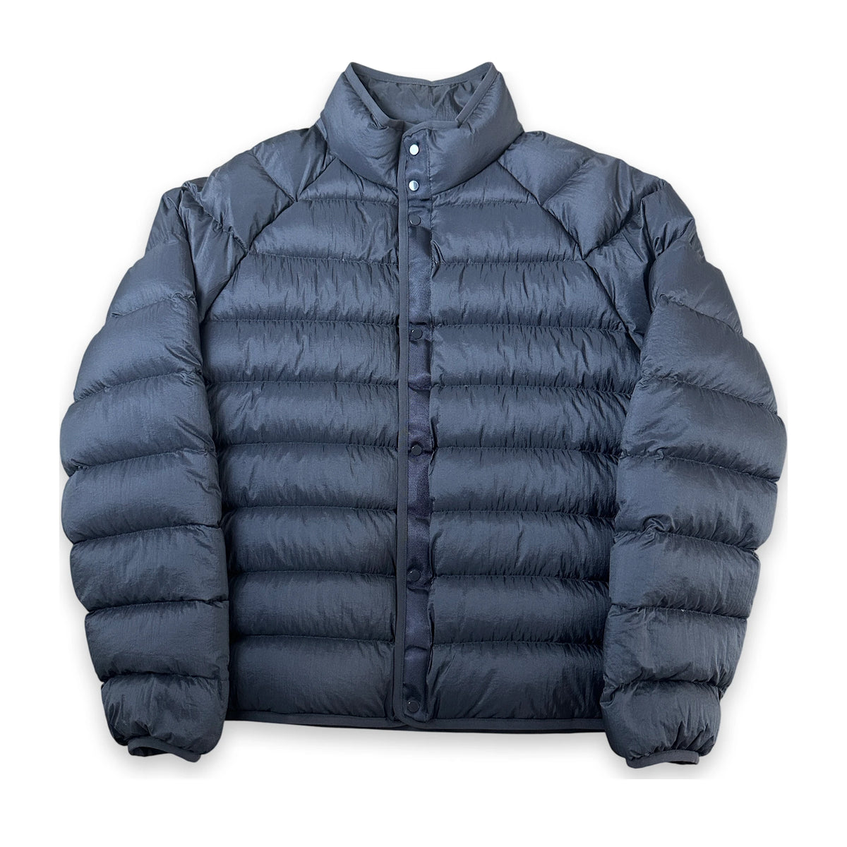 A dark slate blue puffer jacket with horizontal quilting, a stand-up collar, and a snap-button front closure, displayed against a solid white background.