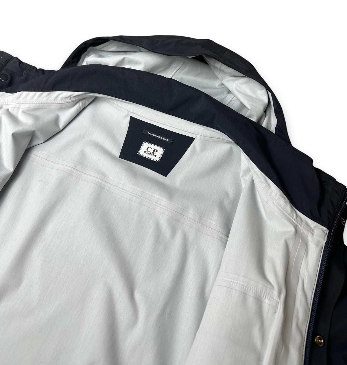 Close-up of a jacket interior showing a light gray fabric body, dark navy collar and hood lining, and a small interior label with CP branding.
