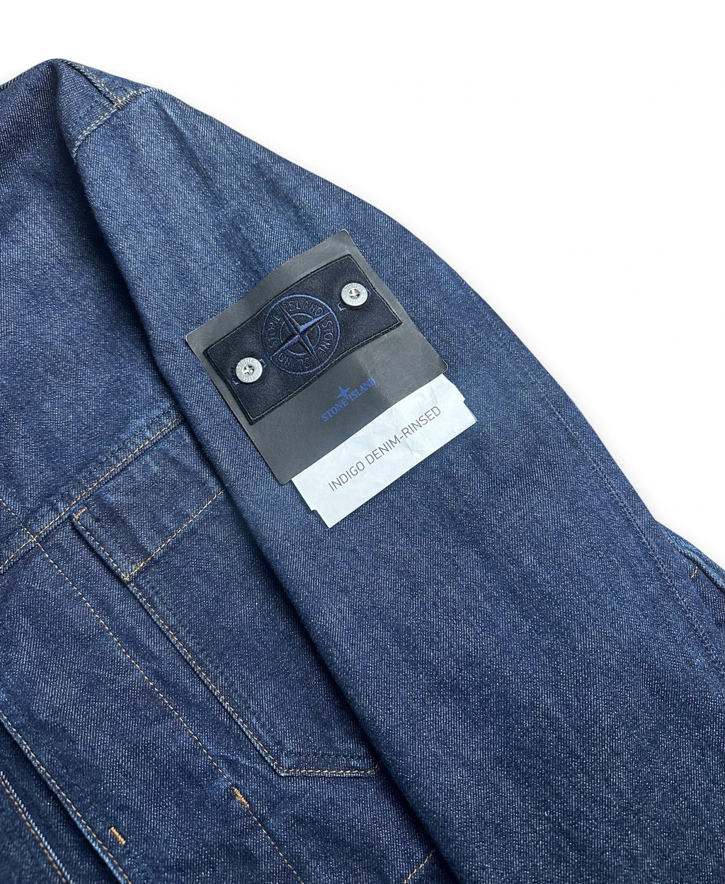 Close-up of a Stone Island denim jacket in blue denim, showing the brand's signature patch on the chest pocket.