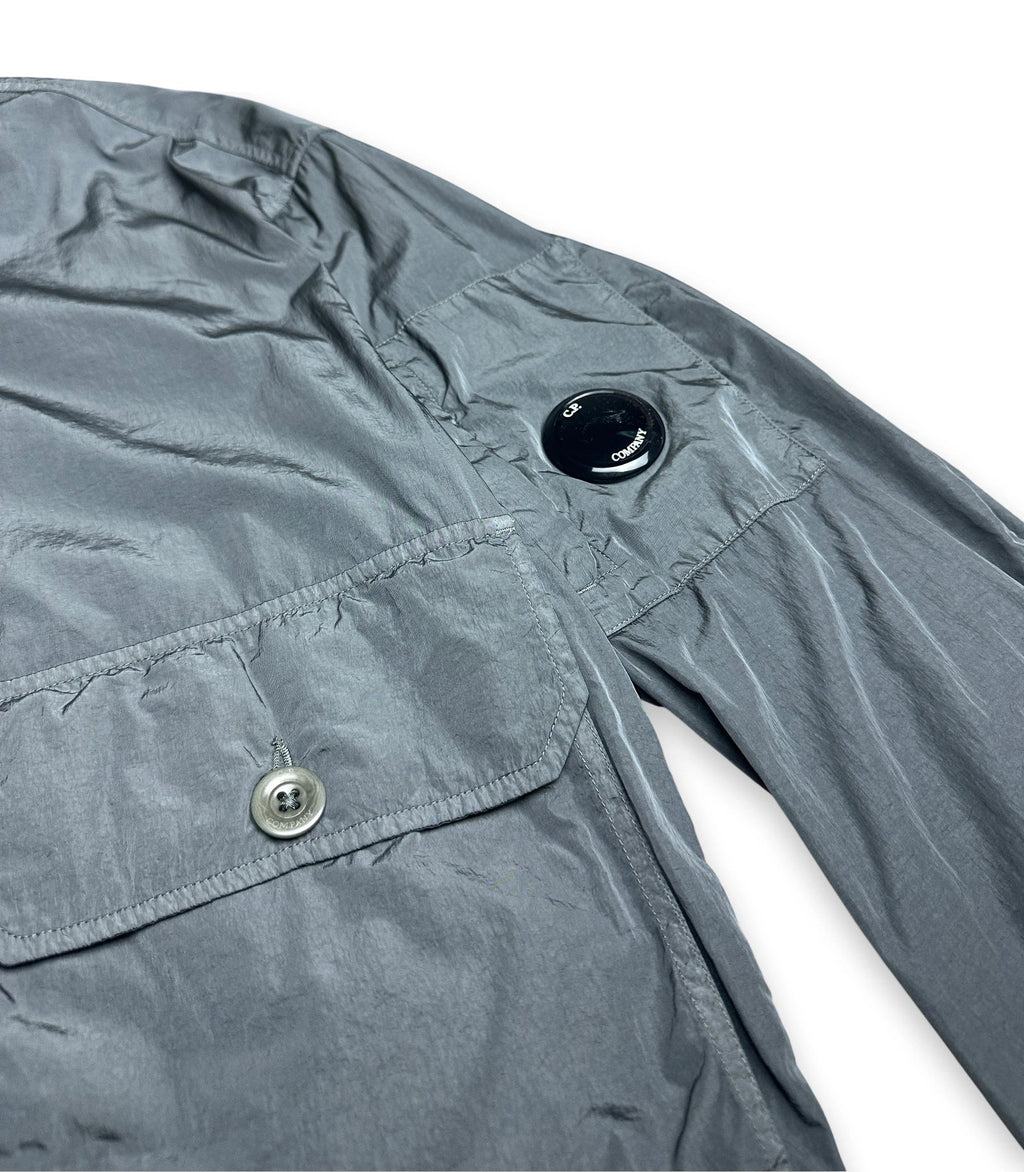 Close-up of a gray lightweight jacket or overshirt showing part of the sleeve, a chest pocket with a silver button, and a black circular shoulder detail on a white background.
