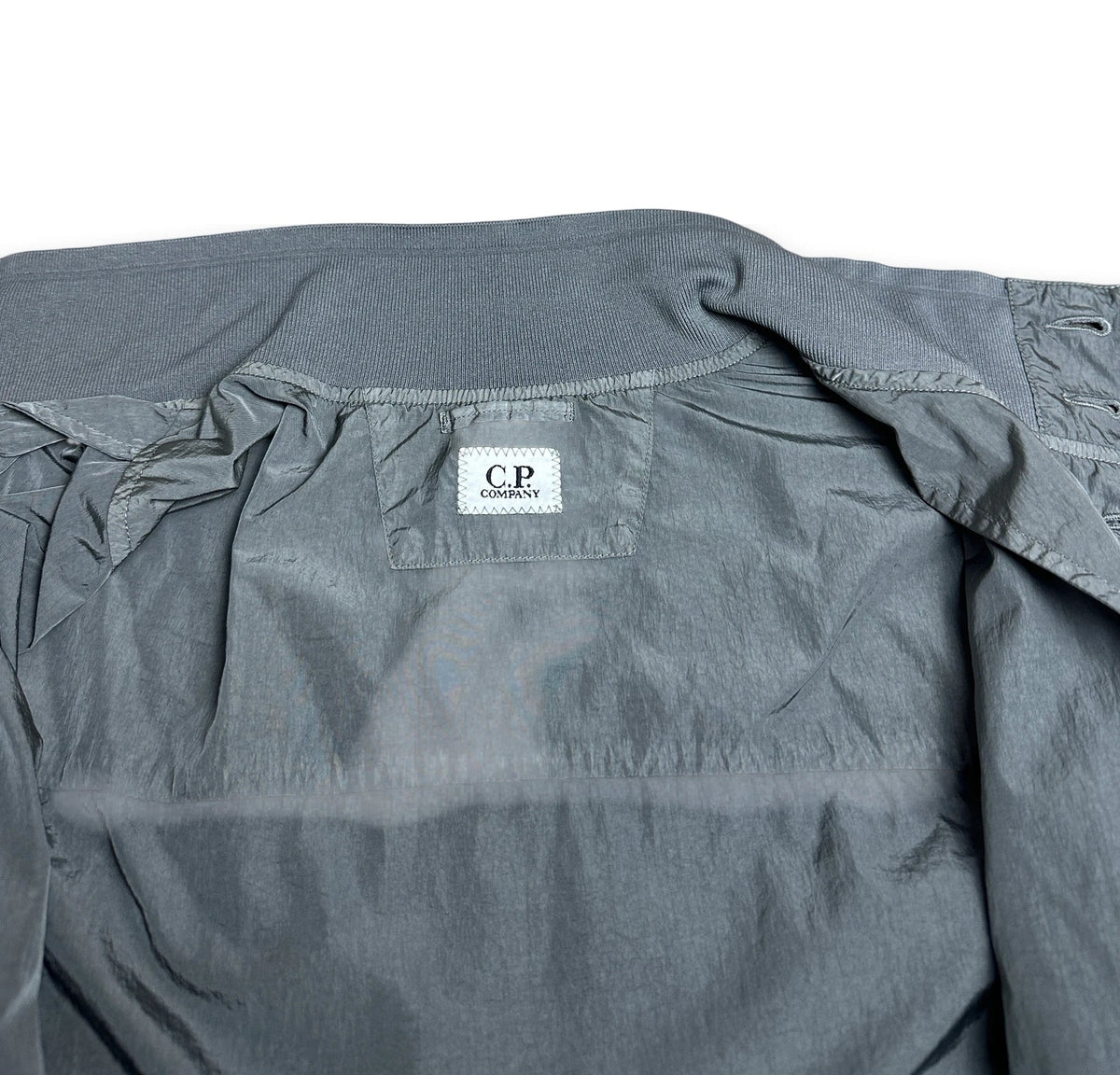 A close-up of a gray lightweight jacket or windbreaker, showing the inner collar area with a white brand label that reads 'C.P.