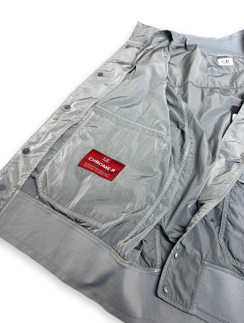 A close-up of a light gray metallic jacket or overshirt laid flat on a white background, showing a chest pocket with a red rectangular label that reads 'CHROME-R'.