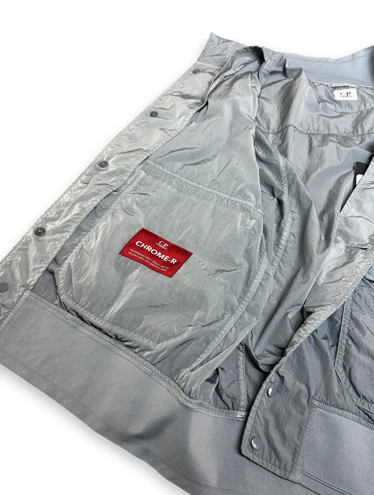 A close-up of a light gray metallic jacket or overshirt laid flat on a white background, showing a chest pocket with a red rectangular label that reads 'CHROME-R'.