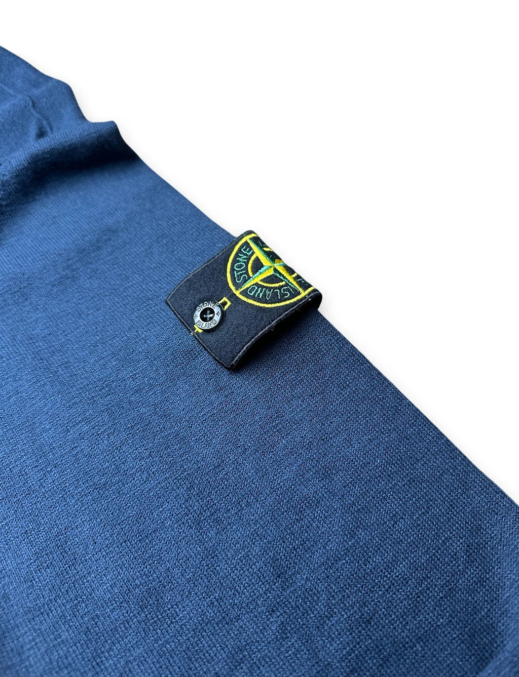 Close-up of a blue knit garment with a black Stone Island brand tag sewn onto the shoulder, featuring the iconic compass logo.