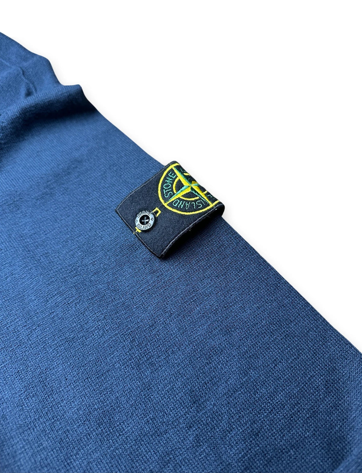 Close-up of a blue knit garment with a black Stone Island brand tag sewn onto the shoulder, featuring the iconic compass logo.
