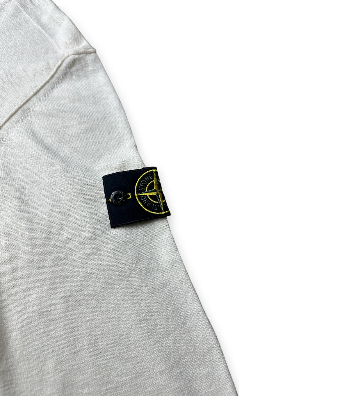 Close-up of a light gray heathered t-shirt sleeve featuring the black Stone Island brand tag with its signature yellow compass logo.