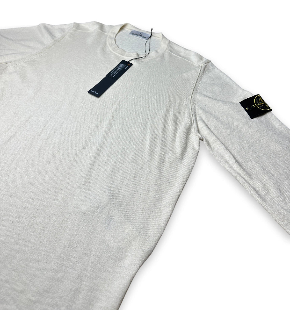 A long-sleeve crewneck shirt in off-white/cream, featuring the black Stone Island compass patch on the left sleeve and a dark blue hangtag near the collar.