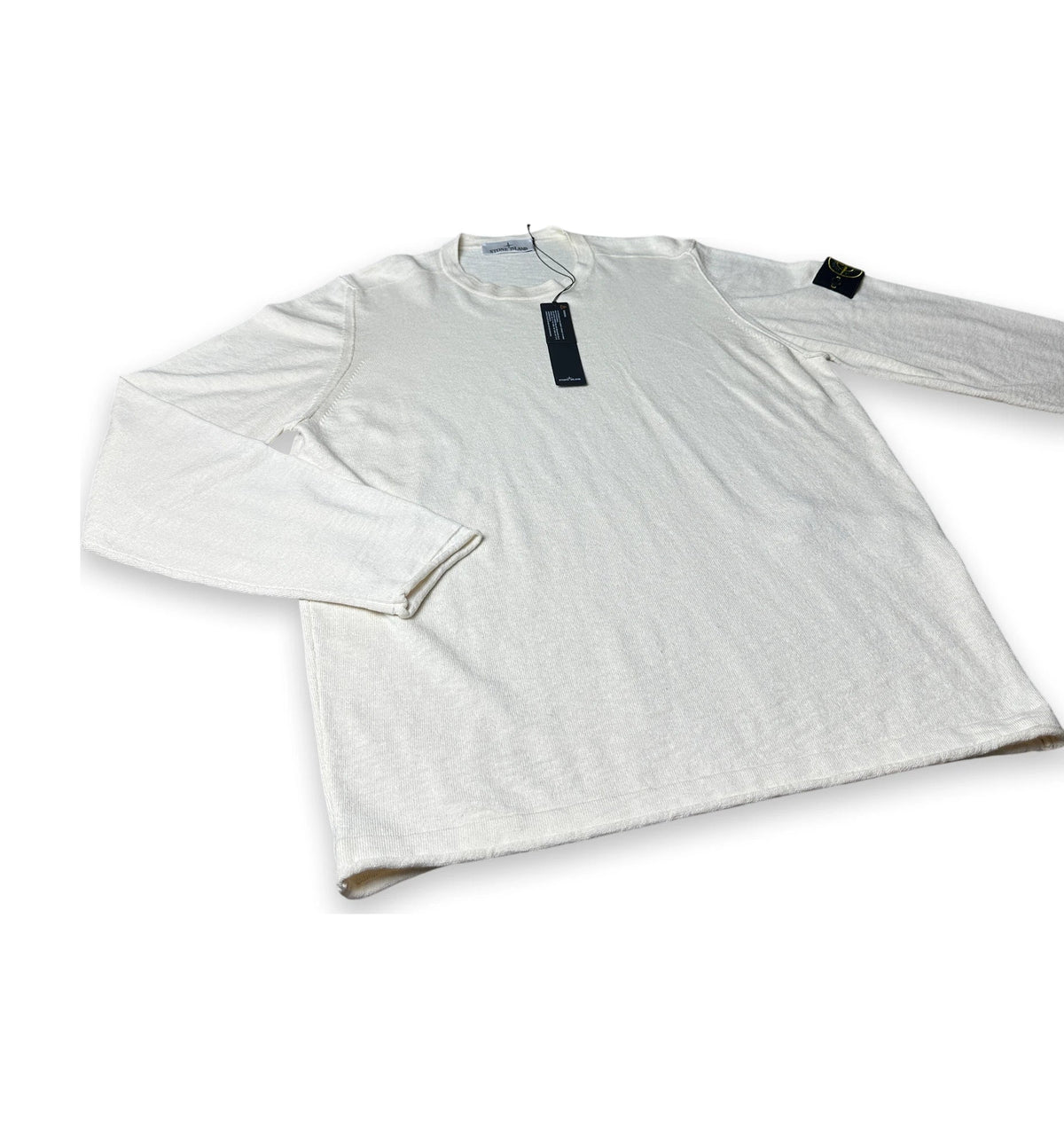 A long-sleeve crewneck shirt in off-white or cream, laid flat on a white background. It features a black hangtag at the collar and a small black rectangular patch on the upper left sleeve.