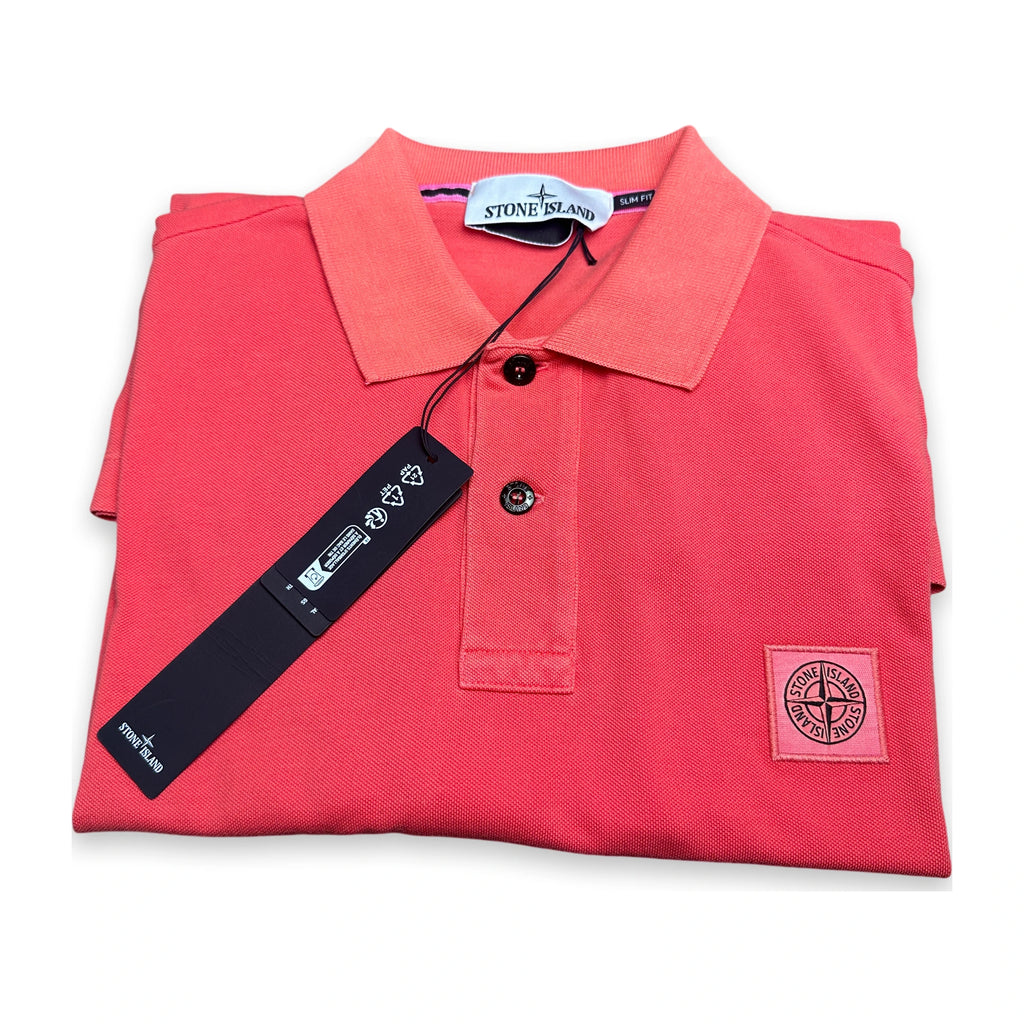 A folded coral pink Stone Island polo shirt with its black hangtag and brand label visible, set against a plain white background.