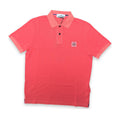 A coral pink short-sleeve polo shirt with a three-button placket and a small circular compass logo patch on the left chest, displayed flat against a white background.