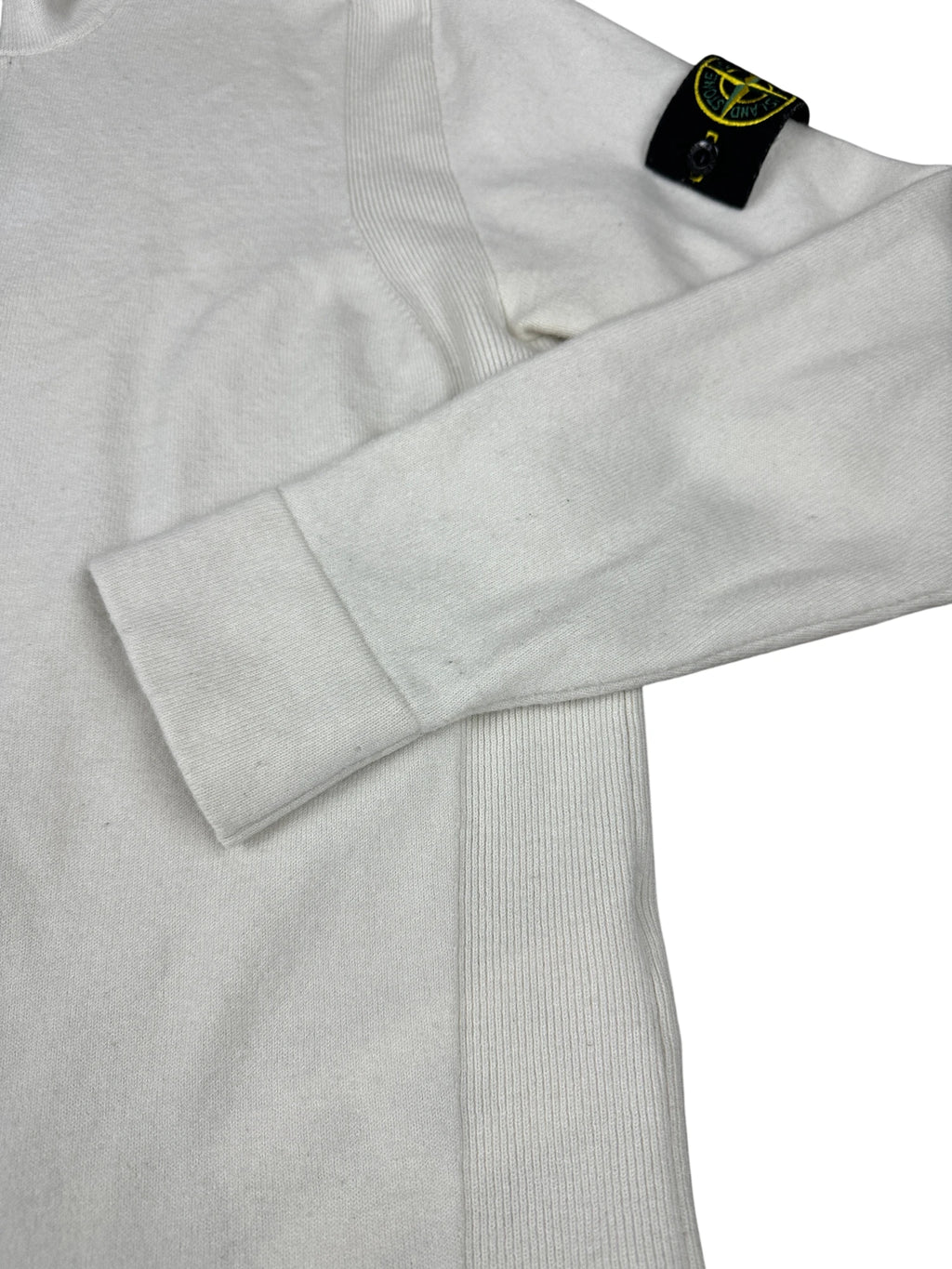 A close-up of a light gray ribbed knit sweater with a folded cuff and a black Stone Island neck label featuring the yellow compass logo.