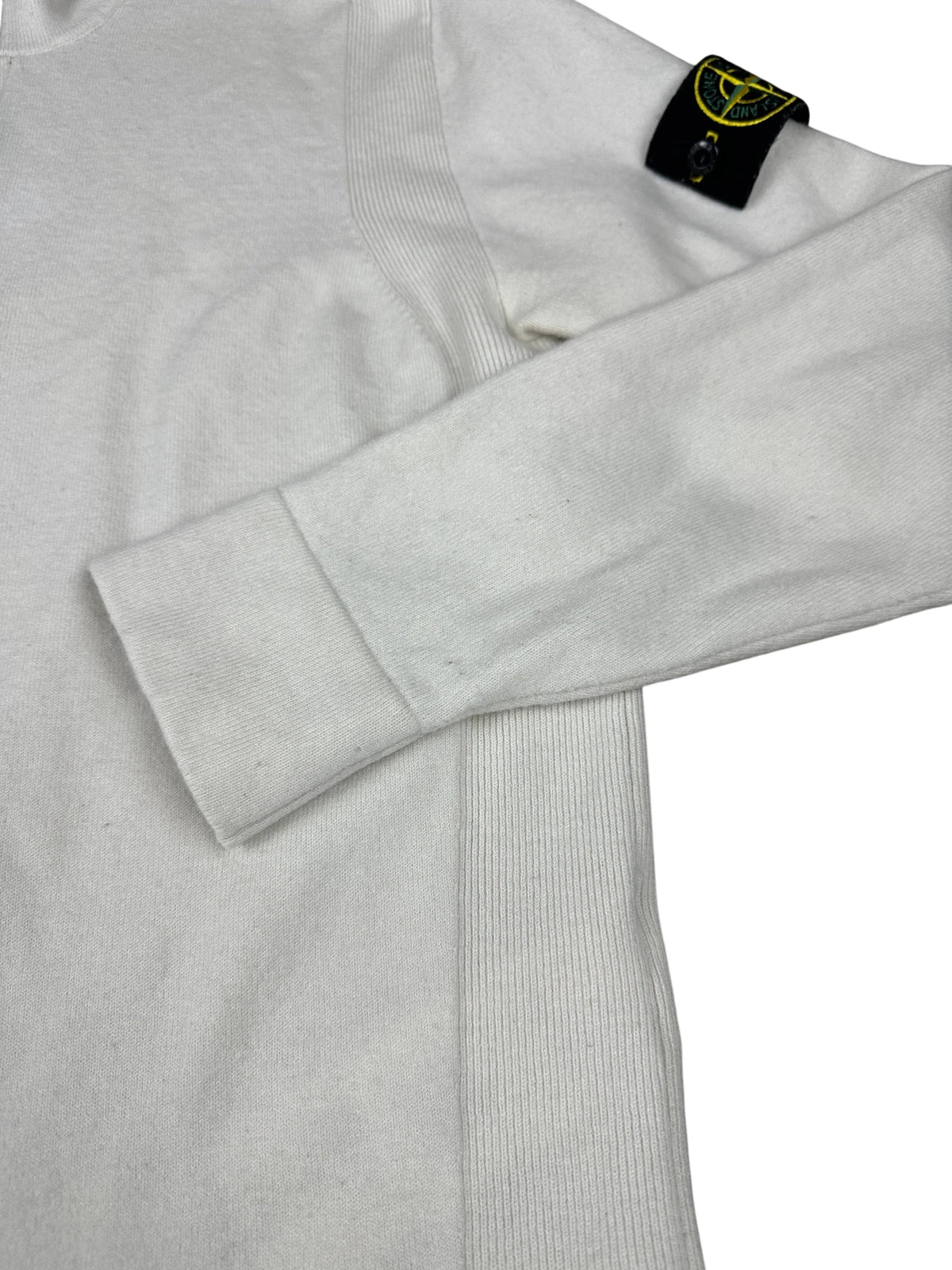 A close-up of a light gray ribbed knit sweater with a folded cuff and a black Stone Island neck label featuring the yellow compass logo.